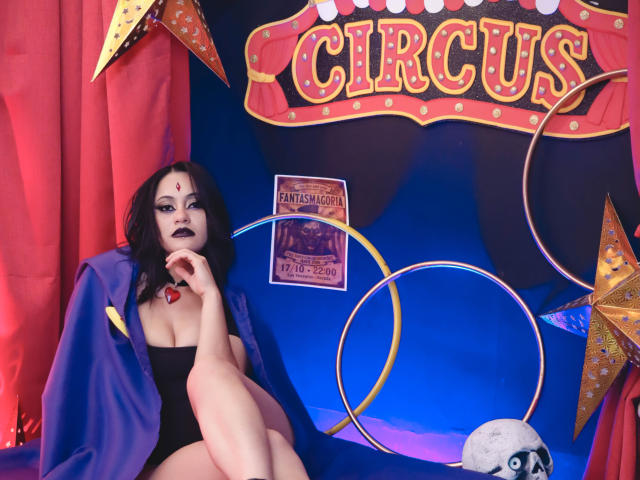 CassieJones with black lipstick, red heart on forehead, black strapless top, purple cape, sitting, showing tits, legs crossed, red curtains, blue backdrop, star and ring props, moon.