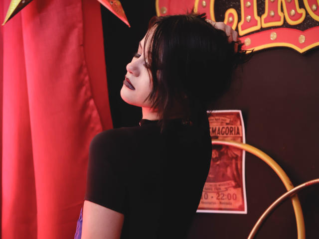 CassieJones with dark lipstick, black shirt, and short black hair, leaning against a red curtain, hand in hair, looking up, showing her ass, in a dimly lit room.
