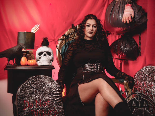 ElizabethDupont with curly dark hair, black lace top, and skirt, sitting on a chair, showing her tits, with a skull, pumpkin, and crow on a red table.