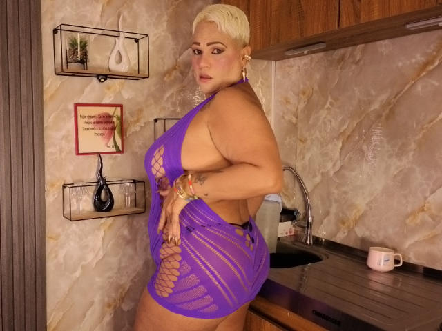 GianellaCox se tient dans une cuisine, vêtue d'une robe violette en dentelle qui révèle ses fesses volumineuses et sa poitrine, avec des cheveux blonds courts, et regarde par-dessus son épaule..