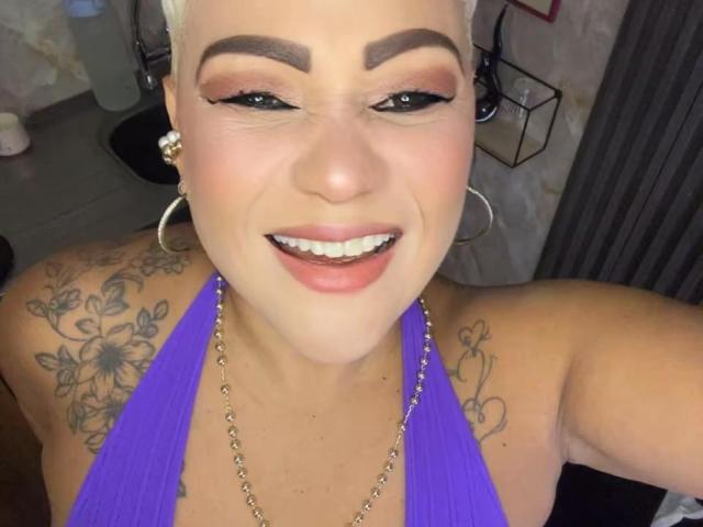 Gianella Cox, avec ses cheveux blonds courts, ses sourcils épais, son eye-liner noir, son haut violet à encolure dégagée, ses boucles d'oreilles et ses tatouages sur les épaules, sourit en montrant ses dents blanches, et sa photo révèle une partie de ses fesses..