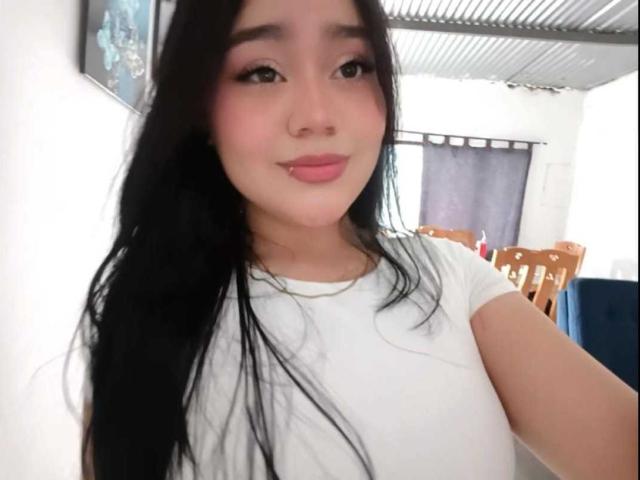 MalloryRamos with long black hair, pink blush, and white t-shirt, smiling at the camera, showing a subtle lip piercing, in a well-lit room with a ceiling vent.