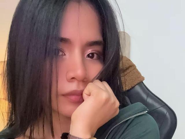 SaschaHelen with dark straight hair, slightly parted lips, and brown eyes, holding her chin with a green jacket, looking forward. She's got a cute face, and she's sexy.