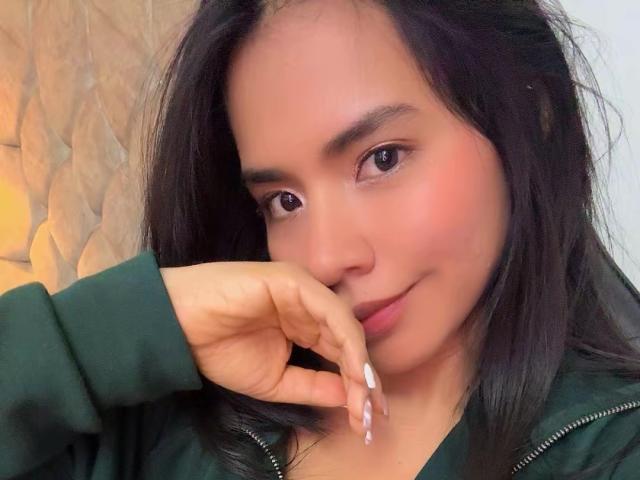SaschaHelen with dark hair, medium skin, brown eyes, slight smile, green jacket, hand with long white nails touching chin, looking at camera, soft pink blush on cheeks.