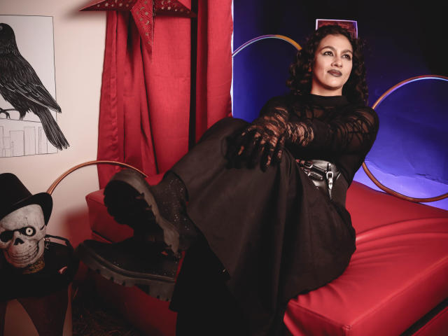 ElizabethDupont with curly dark hair, wearing a black lace dress and gloves, black boots, and a corset, sitting on a red couch, legs up, showing her ass.