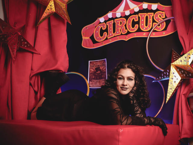 ElizabethDupont with curly black hair, lying on a red bench, wearing a black lace dress, smiling at the camera, surrounded by red curtains and gold star decorations.