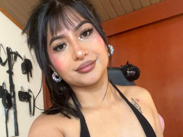 ZarayWhitMoreLove with black hair, bangs, and pink blush, wearing a black halter top, showing small tits, and a tattoo on her right shoulder, smiling at the camera.