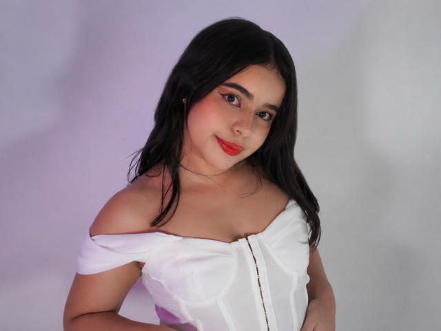 LissyLaurent with black hair, red lipstick, and white off-shoulder top, showing her tits, looking at the camera, smiling. Soft light, subtle makeup, silver necklace.