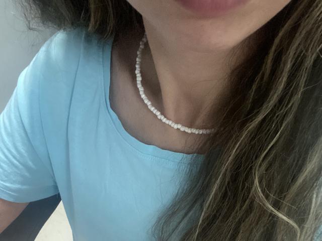 Sanella's face is partially shown, wearing a light blue shirt with a white bead necklace, long wavy brown hair, slightly open lips, soft light on her skin.