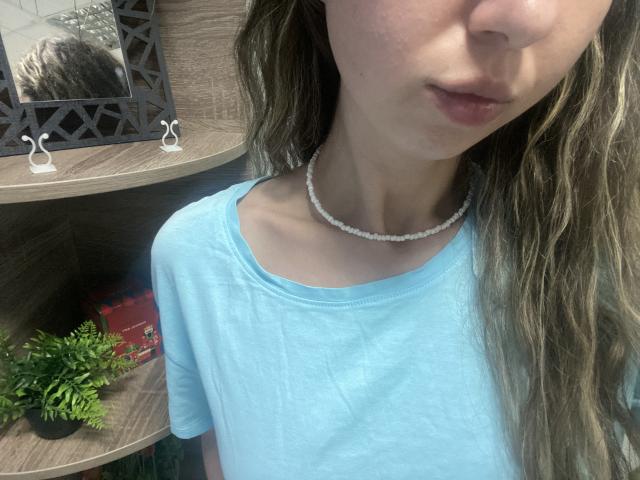 Sanella with light blue shirt, white bead necklace, wavy brown hair, partial face showing lips, small tits, slim neck, wooden shelf with plants and decorative items in background.