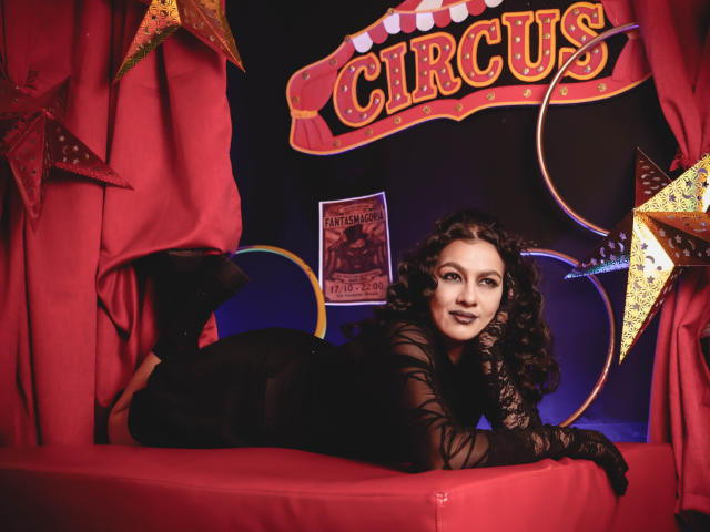 ElizabethDupont with curly dark hair, black lace dress, and black high heels, lying on red stage with stars, looking at camera with a sexy smile, showcasing her ass.