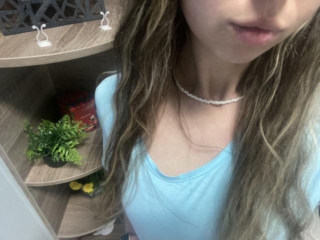 Sanella's face with wavy brown hair, light pink lips, wearing a blue shirt and white pearl necklace, showing a hint of her small tits. Shelves with plants and red car.