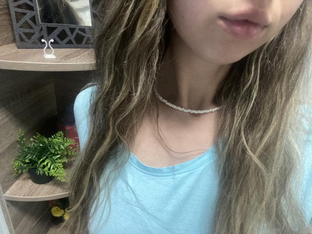 Sanella's face with parted lips, wearing a light blue tee, pearl necklace, and wavy brown hair, showing a slight, cute, and sexy look. Boobs not visible.