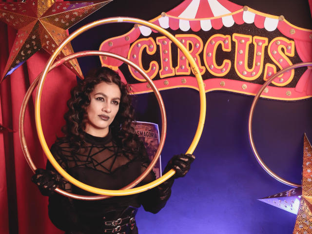 ElizabethDupont with curly dark hair, wearing a black sheer top, black gloves, and holding two golden hula hoops, standing in front of a circus backdrop with star shapes.