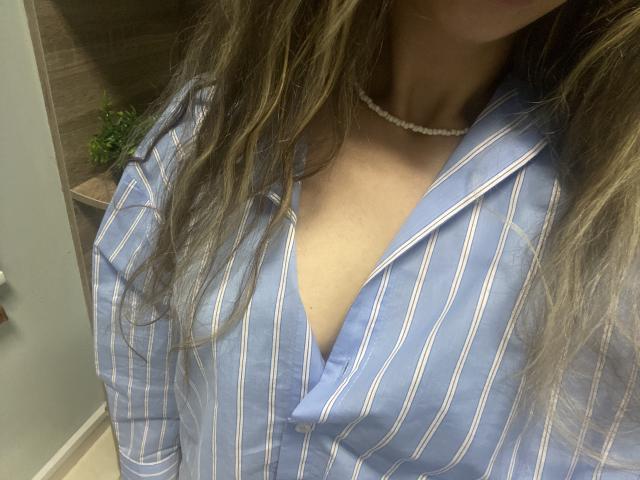 Sanella's blue striped shirt with a deep V-neck, showing a bit of her tits, wearing a pearl necklace, with wavy brown hair, in a dimly lit room.