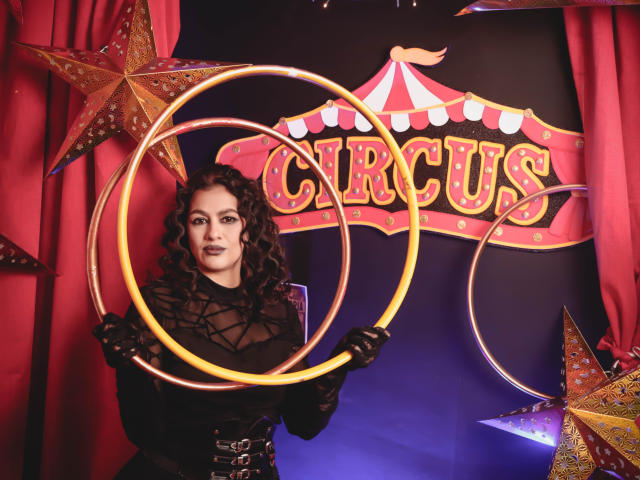 ElizabethDupont with curly dark hair, wearing black lace and corset, holding glowing hula hoops, red star ornaments, and a confident smile, in a circus-themed setup.