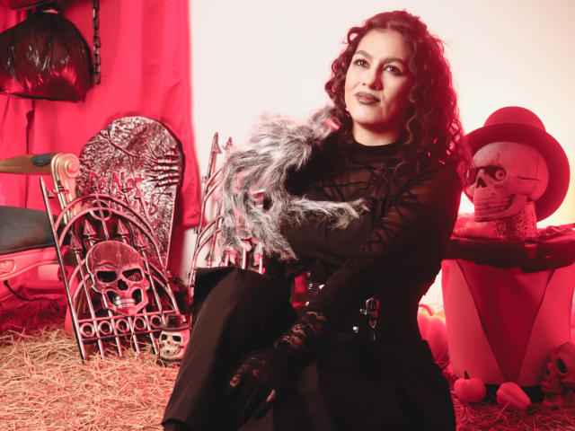 ElizabethDupont with curly dark hair, black lace dress, and gray feather, holding a skull, sitting on hay, surrounded by Halloween decorations, including a skull chair and skeleton.