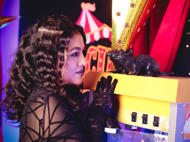 ElizabethDupont with curly black hair, black lace gloves, and geometric black top, staring at a black bat on a yellow box. Red and yellow background. Boobs not visible.