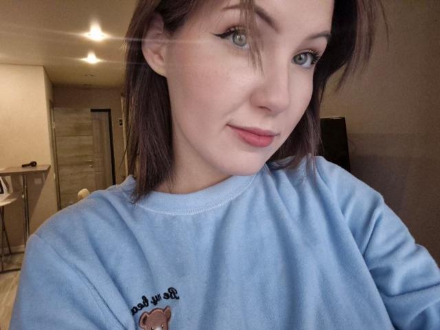 LidiaOckimey has light skin, blue eyes, and brown hair. She's wearing a blue shirt with a small embroidered design, smiling slightly, and looking at the camera.