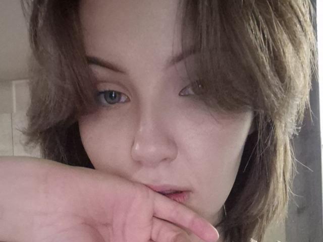 LidiaOckimey has pale skin, brown wavy hair, blue eyes, and a nose piercing. She's biting her finger with a slight pout, looking directly at the camera.