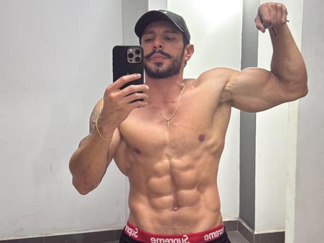 RobertHunk, musclé, torse nu, contractant son biceps, portant une casquette noire, un collier en or, un caleçon rouge avec un logo blanc, tenant un téléphone, barbu, avec un tatouage sur le bras droit, autoportrait pris dans un miroir..