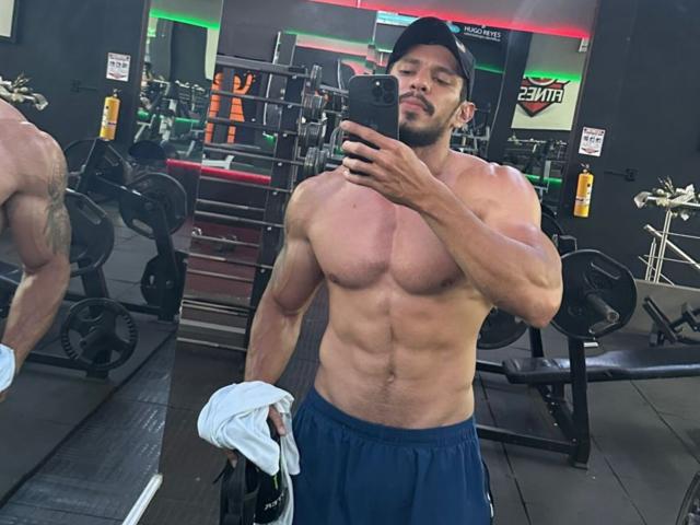 RobertHunk, sem camisa, musculoso, com abdominais definidos, usando um boné preto e shorts azuis, segurando uma toalha branca, tirando uma selfie no espelho em uma academia, mostrando seu traseiro grande..