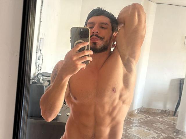 RobertHunk, ohne Hemd, muskulös, mit definierten Bauchmuskeln, schwarzem Haar und Bart, macht ein Selfie vor einem Spiegel, hält ein Telefon, rechter Arm erhoben, Zunge herausgestreckt, steht auf einem braunen Fliesenboden..