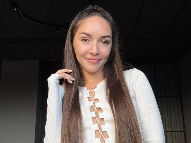 AquaLady with long brown hair, wearing a white lace-up top, smiling slightly, hand near face, soft light, natural look, cute, slim, light skin, relaxed, indoor setting.