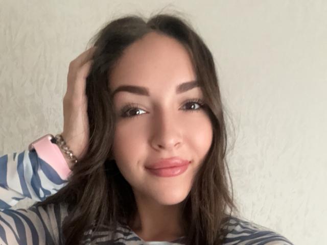 AquaLady with brown wavy hair, pink watch, and striped shirt, hand in hair, smiling with glossy pink lips, soft makeup, and arched eyebrows, looking at camera.
