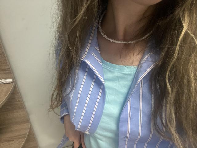 Sanella's blue striped shirt over a light blue dress, with long brown hair and a white bead necklace, showing her upper chest and hand on her hip.