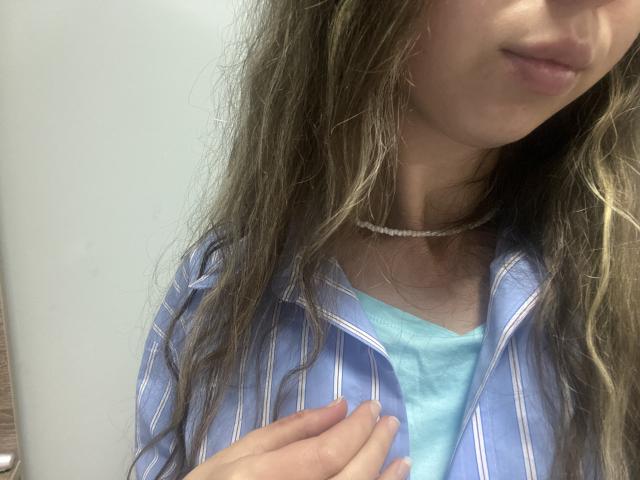 Sanella with long, wavy brown hair, wearing a blue striped shirt over a light blue top, and a beaded choker. Her hand rests on her chest, slightly covering tits.