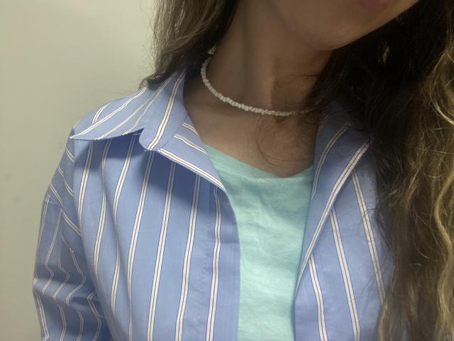 Sanella's blue striped shirt, open to a light blue tee, with a white pearl necklace, showing her neck and upper chest, long brown hair, no visible boobs or tits.