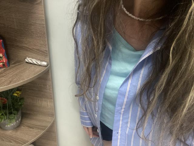 Sanella has long, wavy brown hair, wearing a blue striped shirt over a light blue tank top, and a black thong, with a pearl necklace. Her ass is visible.