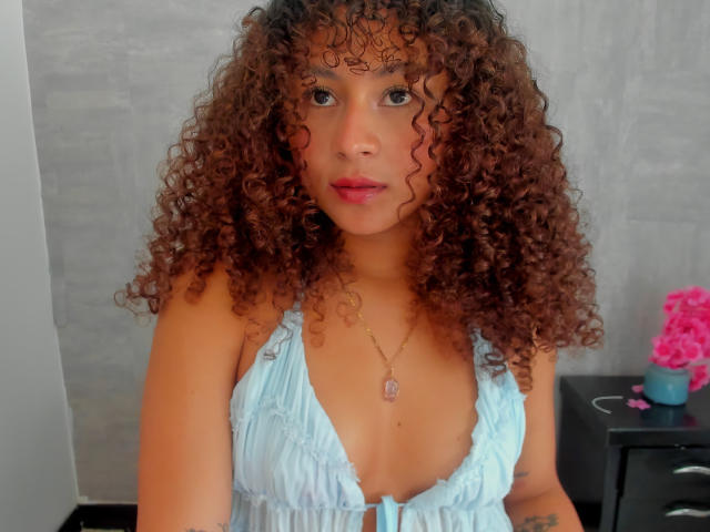 SofiaPassionne with curly brown hair, light brown skin, blue eyes, and pink lips, wearing a light blue, deep V-neck dress, showing her small tits and a tattoo on her arm.