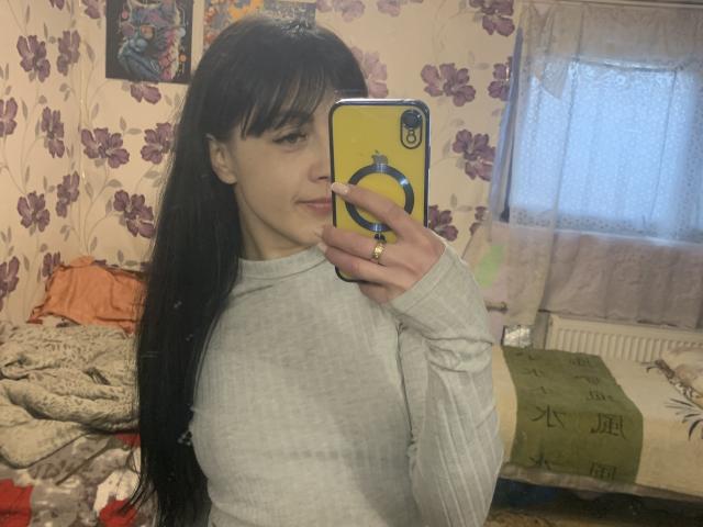 CherryVoss, con lunghi capelli neri, indossa un maglione grigio a coste, tiene in mano un telefono giallo con una cover, sta scattando un selfie allo specchio, mostrando un sorriso appena accennato, in una stanza disordinata..