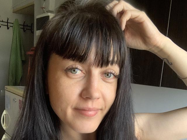 CherryVoss tiene el cabello negro con flequillo, ojos verdes y una ligera sonrisa, sosteniendo su cabello con una mano, mostrando un pequeño tatuaje en su muñeca..