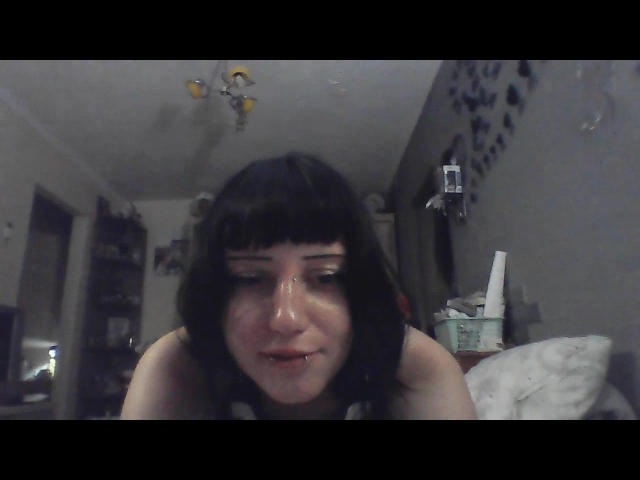 LizaDerrx with black bob, nose dripping with clear liquid, slight smile, lower face and chest visible, looking down, in dimly lit room, ceiling light above.