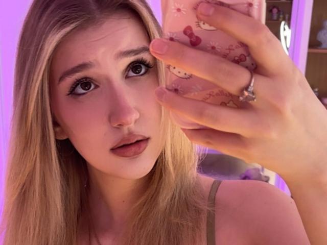Synryx mit blonden Haaren, die überrascht aussieht, hält ein Telefon mit einer rosa Hülle, hat eine rosa Nase und trägt einen Ring am Finger, sowie einen grünen Armband..