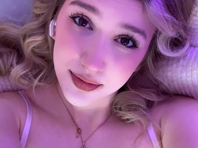 Synryx tiene el cabello rubio ondulado, piel con un tono púrpura, y está mirando a la cámara con una ligera sonrisa, vistiendo una blusa rosa con tirantes finos y un collar. Sus pechos son pequeños..