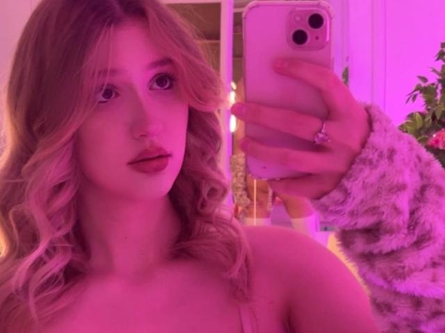 Synryx with pink-tinted hair, wavy locks, holding a phone, wearing a furry sleeve, taking a mirror selfie, showing bare shoulders, pink lipstick, and a ring on her finger.