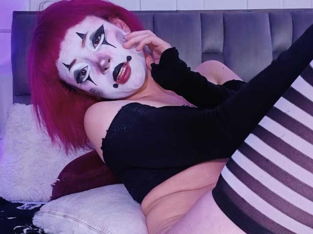ViolettClay with red hair, white clown makeup, black lipstick, and striped thigh-highs, lying on a bed, black off-shoulder top, hand on cheek, showing tits and ass.