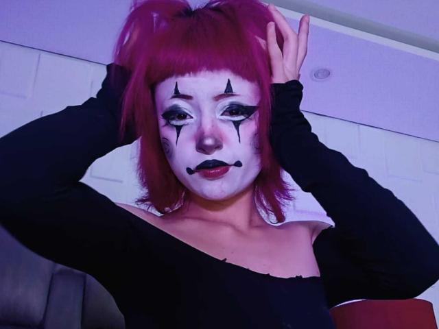 ViolettClay with red hair, white face paint, black eye makeup, and black lipstick, wearing a black off-shoulder top, hands in hair, looking at camera, purple lighting.