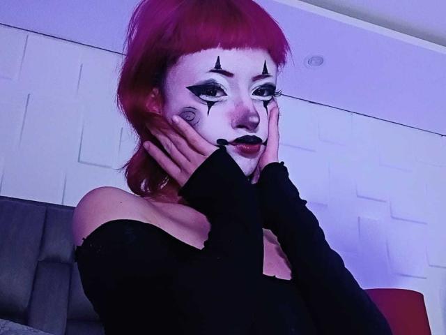 ViolettClay with red hair, black off-shoulder top, and face paint with black eyeliner and lipstick, hands on cheeks, looking at camera, purple-blue lighting.