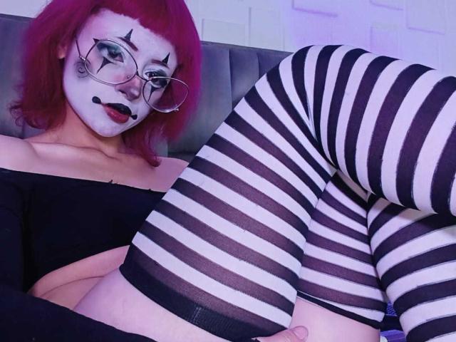ViolettClay with red hair, white face paint, and black lipstick, wearing glasses, black off-shoulder top, and striped black-and-white thigh-highs, showing her ass and a flaccid, circumcised dick.