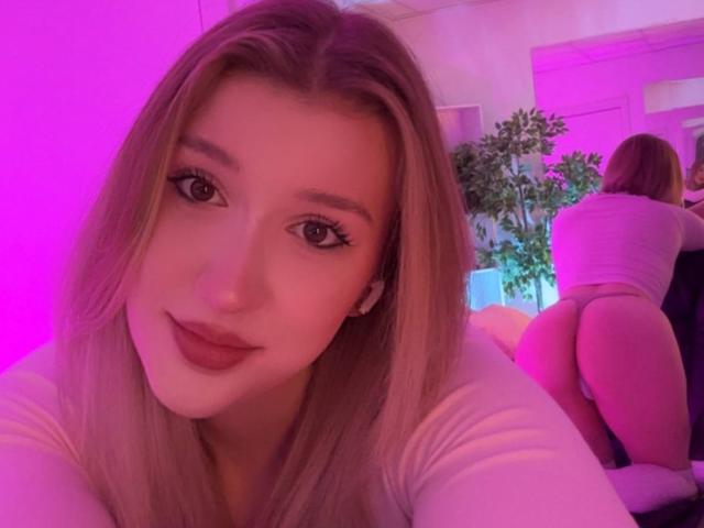 Synryx, avec de longs cheveux blonds et un eye-liner rose, souriant à la caméra, portant une chemise blanche. Ses fesses sont visibles et légèrement relevées en arrière-plan. Éclairage rose et violet..