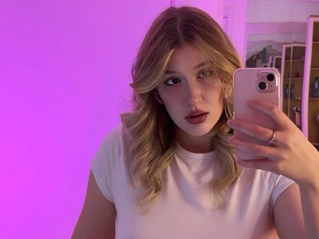Synryx with blonde hair, pink lipstick, and white shirt, taking a mirror selfie with a pink-lit background, holding a pink iPhone, showing a ring on her finger.
