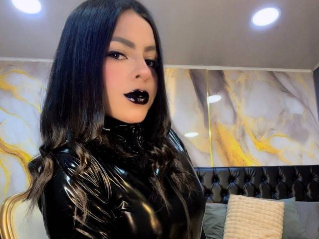 LindaFrancoo tem cabelos longos e pretos, veste uma blusa preta de látex brilhante, com batom preto e maquiagem escura nos olhos, olhando para a câmera com uma expressão ligeiramente satisfeita..