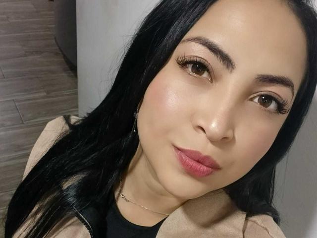 AlanaWest has black hair, brown eyes, and pink lipstick. She's wearing a beige jacket over a black top, with a thin necklace. Her tits are not visible.