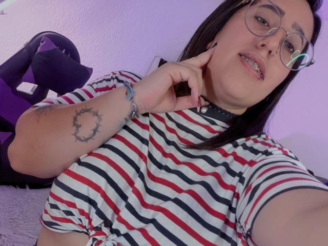 Natalie Harris, usando óculos, com um piercing no nariz e um piercing no septo, vestindo uma camisa listrada, mostrando uma tatuagem no braço e um bracelete, posando com o dedo perto da boca..