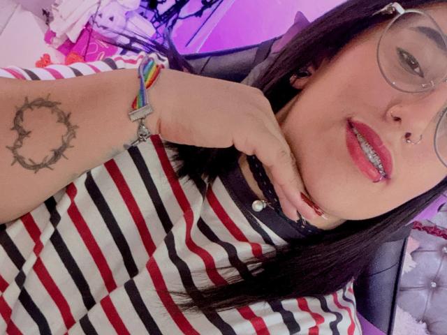 Natalie Harris, com cabelo escuro, óculos, piercing no nariz e no lábio, vestindo uma camisa listrada, uma pulseira colorida e um colar preto, mostrando uma tatuagem de coroa na mão, sorrindo..